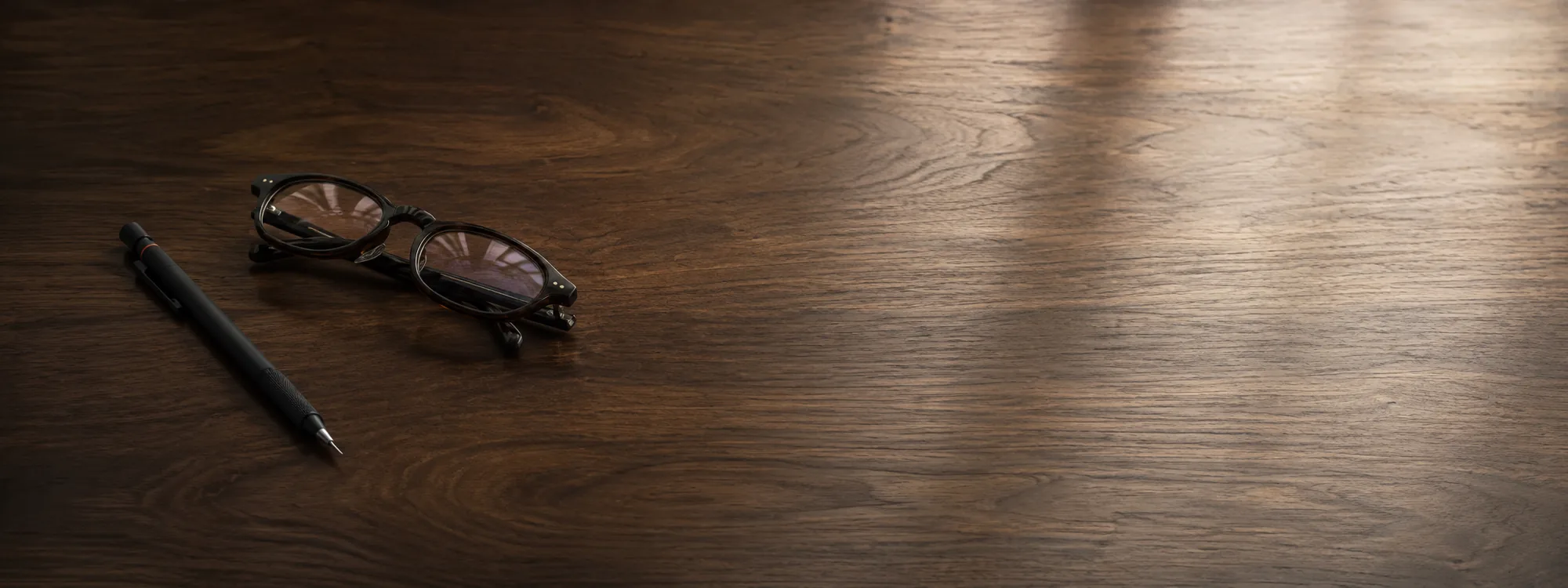 A mechanical pencil and reading glasses on a dark walnut desk